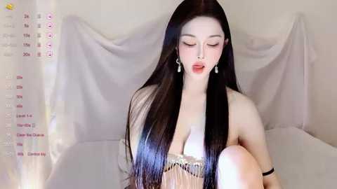 A fair-skinned East Asian woman with long, straight black hair, wearing a sheer, low-cut, beige dress with a silver trim, sits on a bed with white sheets, eyes closed, lips parted, against a soft, draped white background.