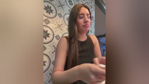 Video of a young woman with long brown hair, fair skin, and glasses, wearing a black tank top, seated at a kitchen counter with tiled backsplash featuring geometric patterns.