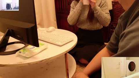 Video of a woman with long hair in a cream sweater and black skirt sitting at a white marble desk with a computer and phone, background includes a red chair and beige wall.
