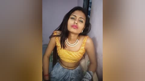 Video of a young South Asian woman with long black hair, wearing a yellow crop top and blue skirt, posing seductively against a plain wall, lips slightly parted.