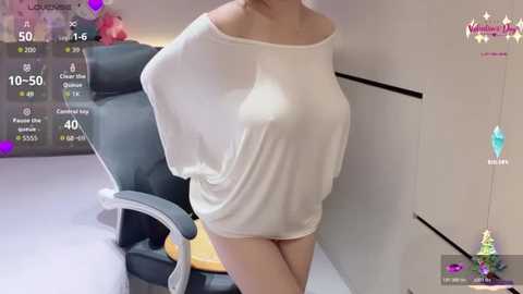 Video of a woman in a sheer white top, revealing her breasts, standing in a modern office with a black chair and white cabinets.