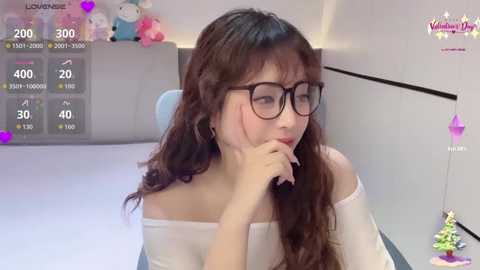 A video of an Asian woman with long brown hair, wearing glasses and a white off-shoulder top, holding a phone, set in a modern, minimalist room with a digital clock and stuffed animals.