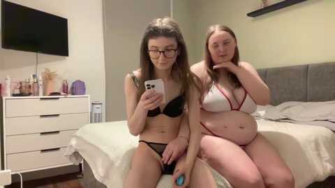 Video of two young women in lingerie, sitting on a bed in a modern bedroom with a TV, dresser, and green walls.