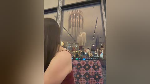 Video of a woman with long brown hair, wearing a red sleeveless top, standing in front of a large window displaying a vibrant, city skyline with skyscrapers and a colorful decorative cloth.