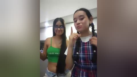Video of two young Asian women in casual attire, one in a green crop top, the other in a plaid shirt, standing in a dimly lit room with a blurred background.