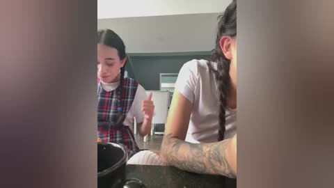 Video of a young woman with dark hair, wearing a plaid dress, and a man with tattoos, both seated at a table in a dimly lit room.