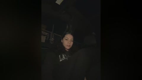 A dimly lit video of a young woman with straight black hair, wearing a black hoodie with \"LA\" on it, taken in a dark room with minimal lighting.