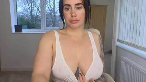 A video of a plus-sized woman with fair skin and dark hair, wearing a low-cut white tank top, sitting indoors near a window with a view of trees.