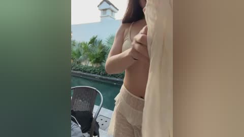 Video of a partially nude woman with medium breasts, holding a white sheet in front of her. She wears beige pants. Background features a pool, greenery, and a white house.