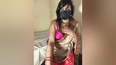 Video of a woman in a pink bra, black mask, and red sari, seated on a bed in a dimly lit room with beige walls.