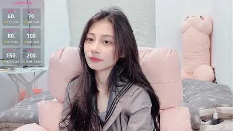 Video of a young Asian woman with long black hair, wearing a light gray robe, sitting on a pastel-colored bed in a cozy, softly lit bedroom.