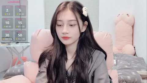 A video of an East Asian woman with long black hair, wearing a black top, seated in a pink, plush, cushioned room with a wall calendar and plush toys.