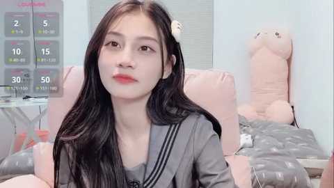 Video of an East Asian woman with long black hair, wearing a grey sailor uniform, sitting on a pink cushioned chair in a minimalist room with pastel-colored decor.