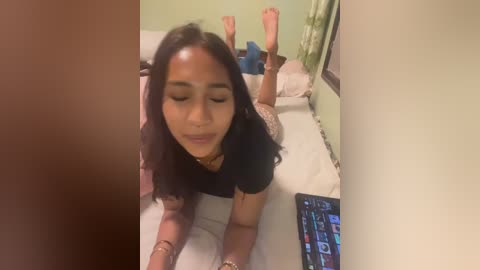 Video of a young woman with long dark hair lying on her stomach on a bed, wearing a black top, playing a video game with her feet up, in a dimly lit room.