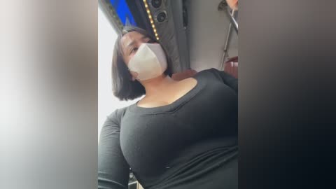 Video of a woman with medium skin tone, shoulder-length black hair, wearing a black long-sleeve top and white face mask, seated in an airplane with overhead compartment visible.