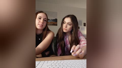 A video of two young women, one with long brown hair in a black top, the other with long brown hair in a tie-dye shirt, sitting at a desk, with blurred faces in the foreground.