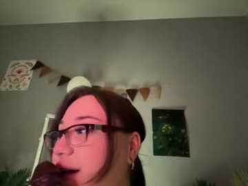 A video of a woman with glasses and a dark headband, standing indoors with a gray wall, a small window, and a decorative tapestry.