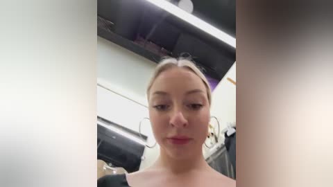 Media: Video of a young, light-skinned, blonde woman with fair skin, wearing a black top, standing in a modern, brightly lit kitchen with stainless steel appliances and white walls.