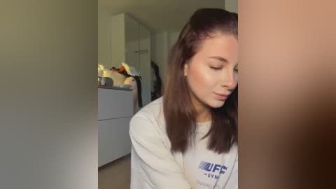 Video of a young woman with straight brown hair, wearing a white long-sleeved shirt, leaning against a wall in a dimly lit, modern bathroom.