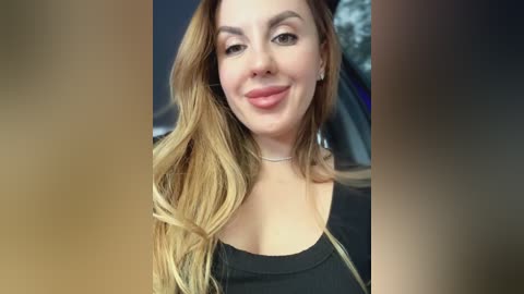 Video of a smiling woman with long, wavy blonde hair, wearing a black top. She has fair skin, a slender build, and is indoors with blurred background.