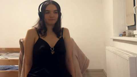 Video of a young woman with long dark hair, wearing black lingerie and headphones, sitting on a bed in a simple, white-walled room with a radiator and a wooden chair.