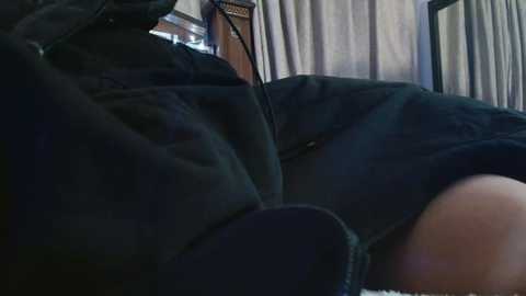 Video of a person lying on a couch, wearing dark clothing, with a blurred, beige curtain in the background. The image is dimly lit, suggesting a cozy, intimate setting.