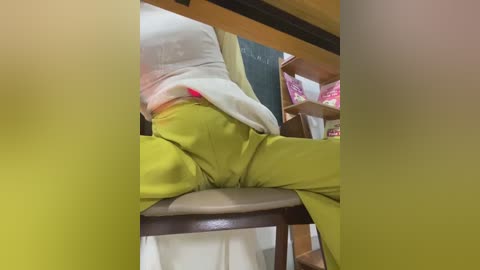 Video of a person wearing a white shirt and bright yellow pants, seated on a wooden chair in a classroom with a chalkboard and bookshelves in the background.