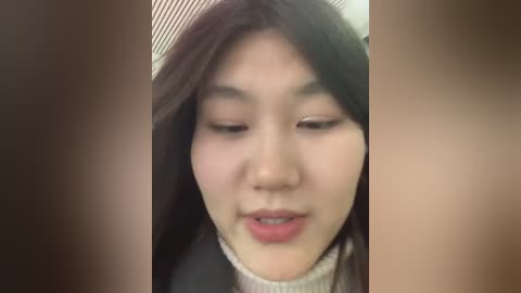 A close-up video of an East Asian woman with long, dark hair, wearing a beige turtleneck sweater, her face partially obscured by blurred, dark-haired figures in the foreground.