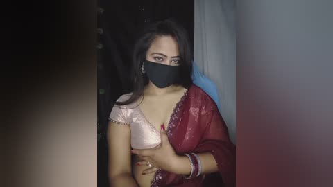 A video shows a woman with long dark hair and medium skin tone, wearing a black mask, red saree, and a pink bralette, standing in a dimly lit room.