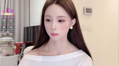 Media: Video of a young East Asian woman with long brown hair, wearing a white off-shoulder top, standing in a modern indoor setting with a glass water dispenser, light beige walls, and a thermostat.