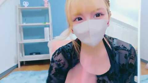 Video of a blonde woman with light skin, wearing a black lace top and a white face mask, sitting on a blue carpet in a brightly lit room with white shelves and a white bed in the background.