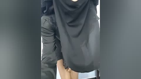 A video of a person wearing a black, long-sleeved, loose-fitting dress with a wide, draped neckline, and a black scarf partially covering their face. The background is dark and blurred, focusing attention on the attire.