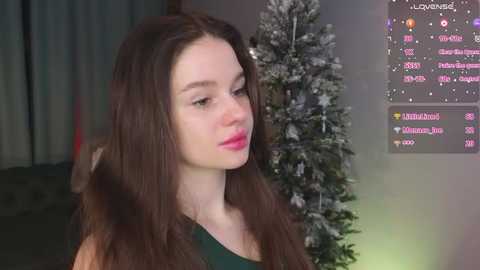 Video of a young woman with long brown hair, fair skin, and pink lipstick, wearing a dark green top, standing in a dimly lit room with a Christmas tree and a virtual chat window.