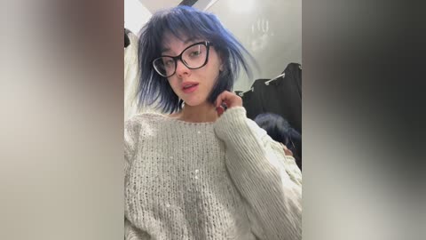 A video of a young woman with short, blue hair, wearing glasses and a cream-colored knitted sweater, posing indoors with blurred background.