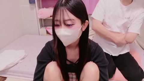 A video of a young Asian woman with long black hair, wearing a white face mask, sitting on a bed in a small, tidy room with pink bedding and a white wall.