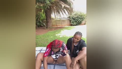 Video of a young African American woman with red braids sitting on a blanket, being assisted by an older woman with braided hair, in a lush garden.