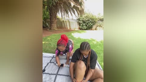 Video of two women with dark skin tones, one with braids, the other with long braids, both wearing black shirts, playing on a grassy lawn with a white and gray checkered blanket.