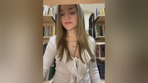 A video of a young Caucasian woman with long, straight light brown hair, wearing a white blouse with a deep V-neck, standing in a library with wooden shelves filled with books.