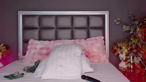 Video of a modern bedroom with a gray tufted headboard, pink and white pillows, a white duvet, and a pair of green glasses on the bed. The walls are painted dark gray, and a glass side table holds a white figurine and a small lamp.
