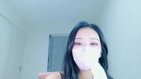 A video of an Asian woman with long black hair, wearing a white face mask, standing in a dimly lit hallway with white walls.