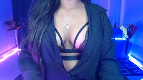 Video of a woman with long black hair, wearing a black lace bra and open shirt, standing in a dimly-lit room with blue LED strip lights.