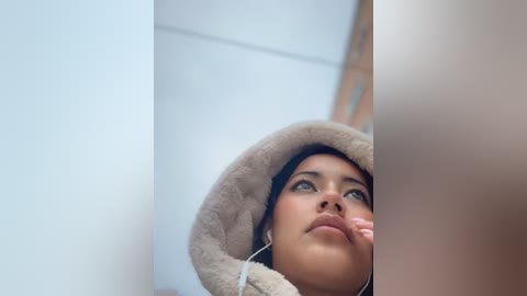 Video of a young woman with olive skin, dark hair, and a light beige fuzzy hood, gazing upward with a contemplative expression. The background is softly blurred, emphasizing her thoughtful demeanor.