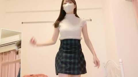 Video of a young Asian woman in a white turtleneck, dark plaid skirt, face mask, and hairband, standing in a bedroom with a closet and white bed visible.