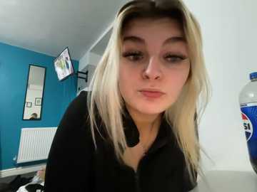 A video of a young woman with long, straight blonde hair and fair skin, wearing a black jacket, leaning forward in a brightly lit room with blue walls, a TV, and a Pepsi bottle on a white table.