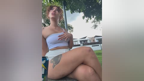 A video of a light-skinned, curly-haired woman in a light blue, off-shoulder crop top and white shorts, sitting on a bench outside, with a tattoo on her thigh.