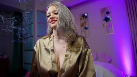 Video of a smiling, fair-skinned woman with long, straight platinum blonde hair wearing a gold satin robe, standing in a softly lit bedroom with purple lighting, decorative wall hangings, and a bed in the background.