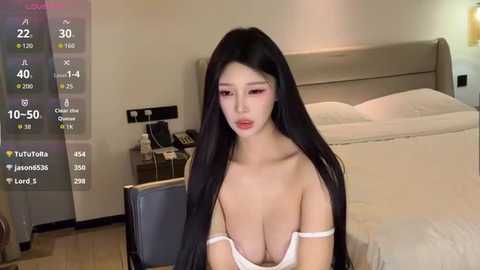 A video of an East Asian woman with long black hair, fair skin, and large breasts, wearing a white bra, sitting in a modern bedroom.