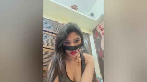 Video of a young Asian woman with long black hair, wearing a black blindfold and red lipstick, standing in a dimly lit room with wooden furniture and decorative items.