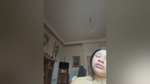 Media: A video of a serene, light-skinned woman with long, straight black hair, wearing a black beanie, lying on a bed in a softly lit, beige-walled room with a chandelier, framed botanical artwork, and a wooden dresser.