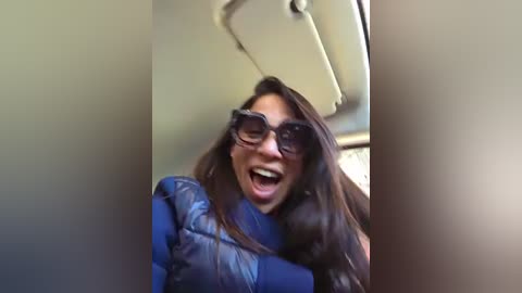 Video of a woman with long brown hair, wearing dark sunglasses and a blue puffy jacket, laughing inside a dimly lit airplane.